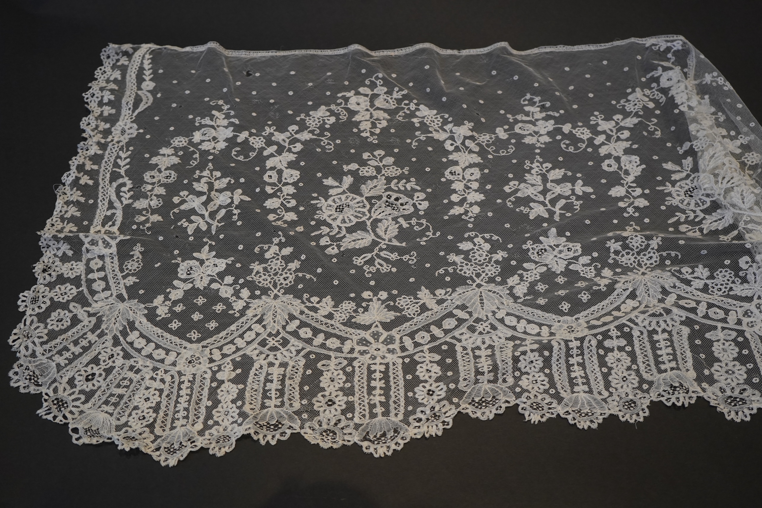 A deep elaborately designed flounce of mid 19th century Brussels bobbin appliquéd lace on net, with finished lace ends, worked with an intricate floral border below large floral cartouches framed in similar motifs and al